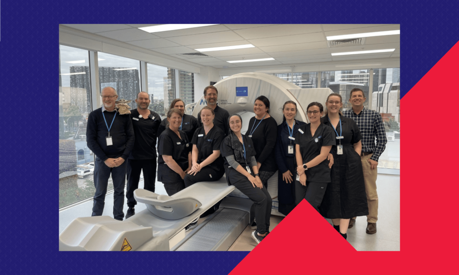 A group of healthcare professionals pose in front of a large medical imaging scanner in a bright, modern facility. The image highlights the successful collaboration and IT infrastructure delivery supporting the operation of the scanner. Tall city buildings are visible through the windows in the background.