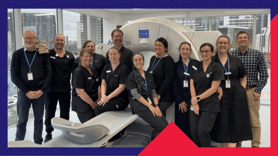 A group of healthcare professionals pose in front of a large medical imaging scanner in a bright, modern facility. The image highlights the successful collaboration and IT infrastructure delivery supporting the operation of the scanner. Tall city buildings are visible through the windows in the background.