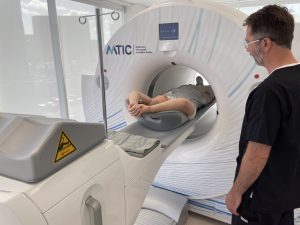 A patient lies on a medical imaging table preparing to enter a CT scanner at the Melbourne Theranostic Innovation Centre (MTIC), while a healthcare professional stands nearby monitoring the procedure. The image showcases the advanced medical technology in use, supported by robust IT infrastructure in a bright, modern clinical setting.